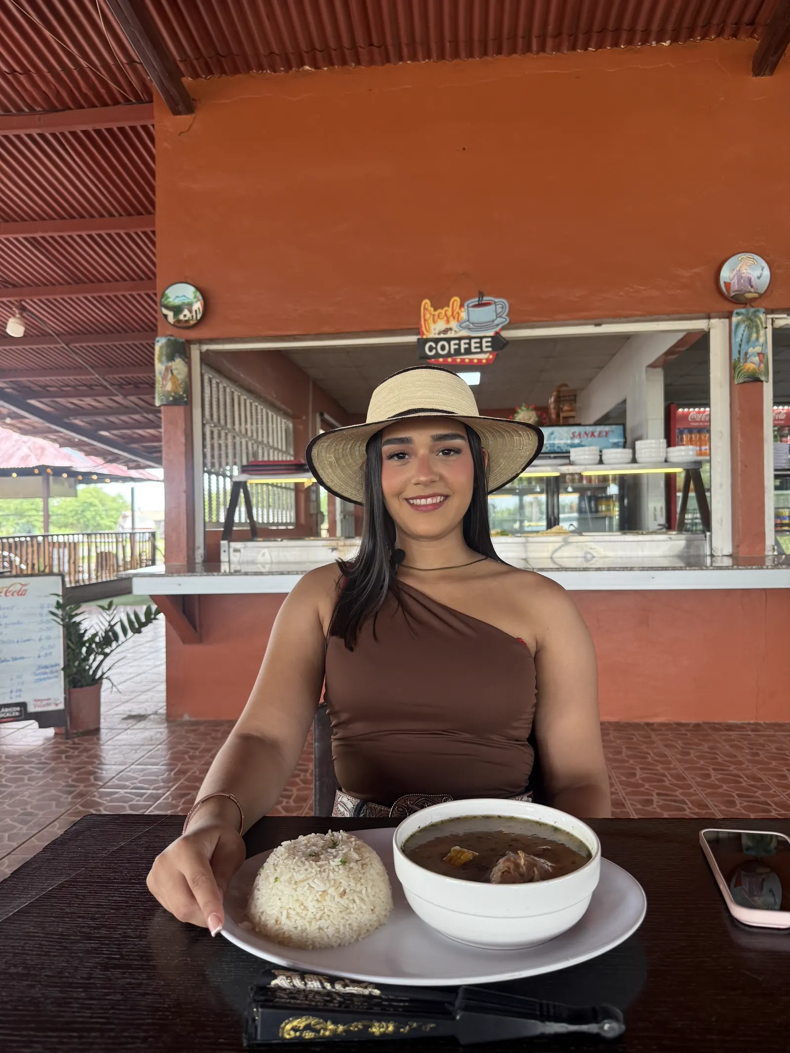 Mujer con sombrero de paja sentada a la mesa con sopa y arroz, frente a la barra de despacho
